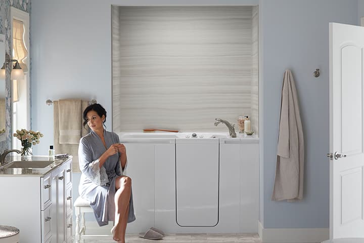 Woman wearing a bath robe sitting in front of her walk in bath tub with her legs crossed and looking towards the vanity sink.