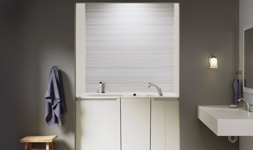 A KOHLER walk-in bath in the cream variation, biscuit, installed in a smaller bathroom with a sconce light on the right beside a sink and mirror and a bath towel and stool on the left.