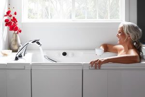Woman sitting in a walk-in tub drinking a cup of coffee while the water is running.