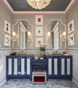 Bathroom with side-by-side blue double vanity with four lit sconces and an overhead chandelier and 2 mirrors above the two sinks and a bench slightly pulled out.