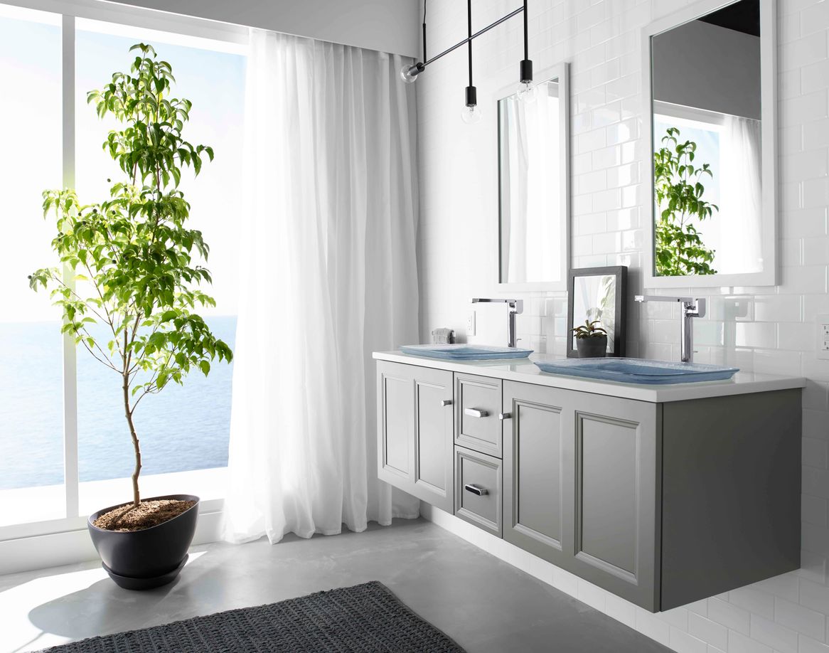 Side view of a bathroom with a gray floating vanity with two sinks and two mirrors above on the right of the image and a tall tree plant in a pot in front of the window to the left of the image.
