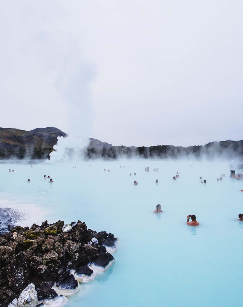 People sitting in the Iceland blue lagoon with mountains in the background and rocks in the foreground