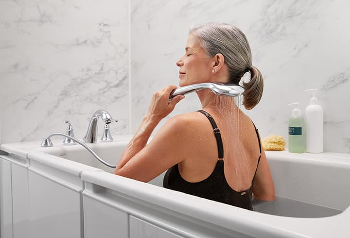 Senior woman wearing a black bathing suit sitting in the walk-in tub with LuxStone bath walls using a handshower to spray her back. 