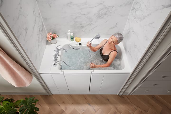 Overhead shot of an older woman wearing a black bathing suit sitting in a jetted walk-in tub using a handshower to spray her chest.
