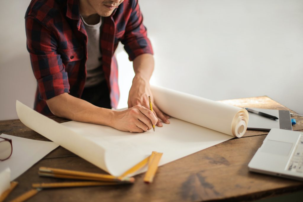 Man bent over a wooden workshop table with a scroll of paper and a wooden pencil in hand