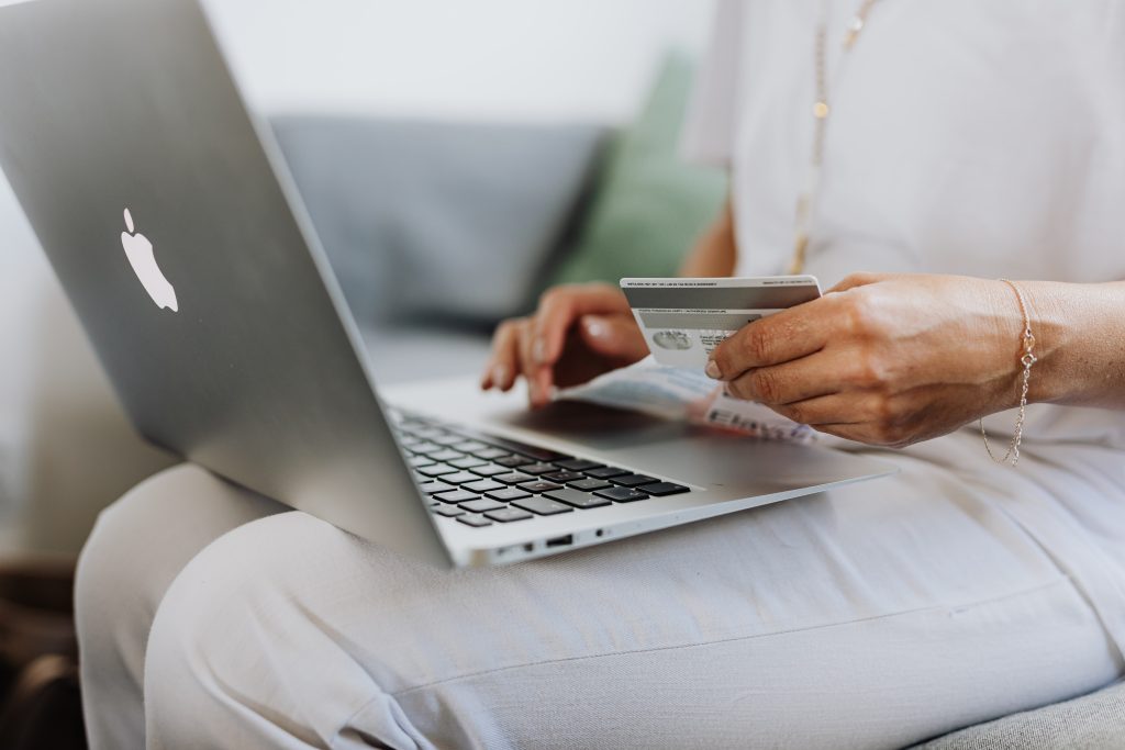 Woman in an all white outfit holding a credit card in one hand and scrolling on a Macbook laptop trackpad with the other hand